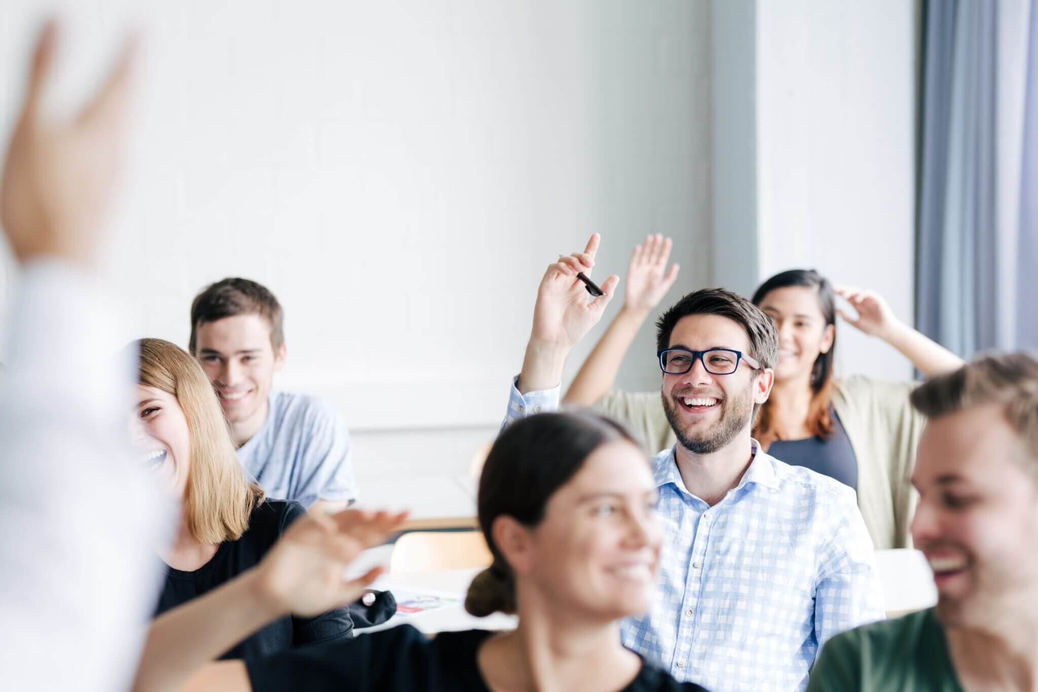 Ein Mann ist im Fokus und meldet sich in einem Kursraum mit anderen Menschen. Der Mann lächelt.